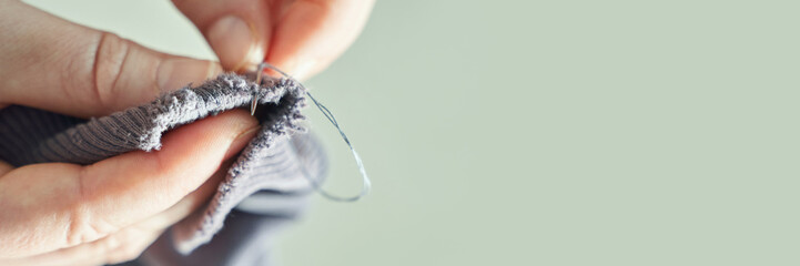 Close-up of hands sewing grey fabric with needle and thread on light green background.
