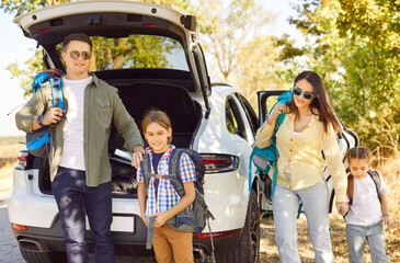 Family with backpacks travel on road trip. Young parents and kids hikers with camping gear walk out...