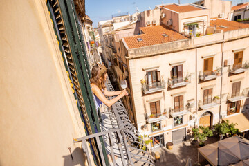 Obraz premium Woman enjoying a peaceful morning on a balcony in Palermo, overlooking old Sicilian buildings under the warm Mediterranean sun. Authentic slow living vibe