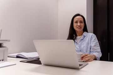 A girl with dark hair in an office with a phone and a laptop at an office desk
