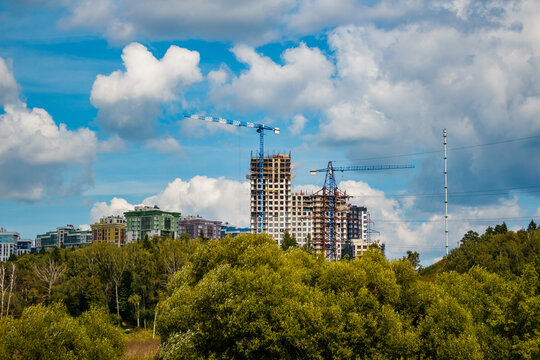 Towering cranes are busy shaping a modern city skyline. New high-rise buildings are under construction, with green trees bordering the urban expansion under a cloudy sky