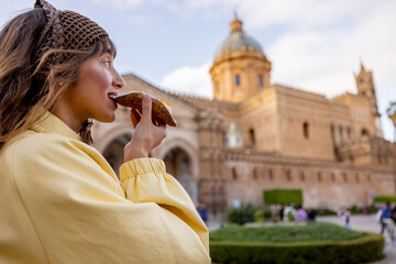 Woman enjoying a bite of traditional Sicilian cannolo in front of Palermo Cathedral. Authentic...