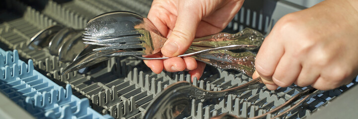 Hands loading silver utensils into dishwasher: forks and spoons in rack.