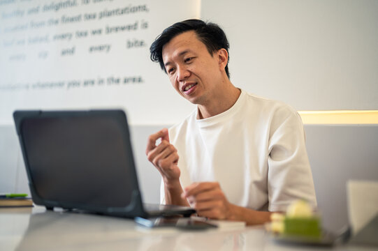 Asian man in a white shirt gesturing with both hands while speaking during an online meeting or discussion at a café table, with a laptop open in front of him in a bright workspace
