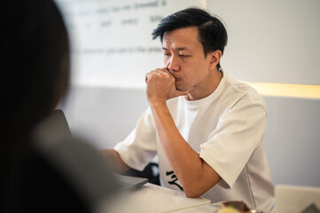 Asian man in a white shirt holding a notebook and pen while thinking deeply at a modern café table, with a laptop in front of him, showing a focused and contemplative expression in a bright setting