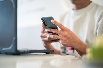 Close-up of an Asian man using a smartphone with both hands while sitting at a café table, a laptop in front of him, wearing a white shirt and a ring, showing focus in a bright workspace