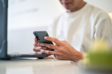 Close-up of an Asian man using a smartphone with both hands while sitting at a café table, a laptop in front of him, wearing a white shirt and a ring, showing focus in a bright workspace