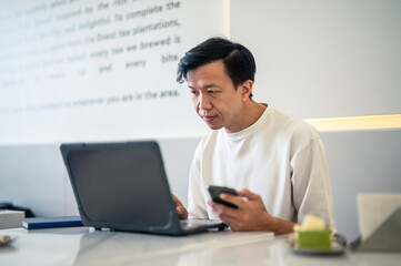 Asian man working at a café table using a laptop while holding a smartphone, appearing focused and engaged in multitasking activities within a modern and bright workspace environment