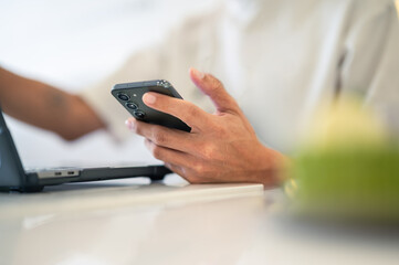 Close-up of an Asian man using a smartphone with both hands while sitting at a café table, a laptop in front of him, wearing a white shirt and a ring, showing focus in a bright workspace
