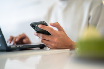 Close-up of an Asian man using a smartphone with both hands while sitting at a café table, a laptop in front of him, wearing a white shirt and a ring, showing focus in a bright workspace