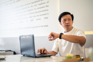 Asian man sitting at a café table using a laptop while checking the time on his wristwatch, showing focus and time management in a bright, modern workspace with a minimalistic background