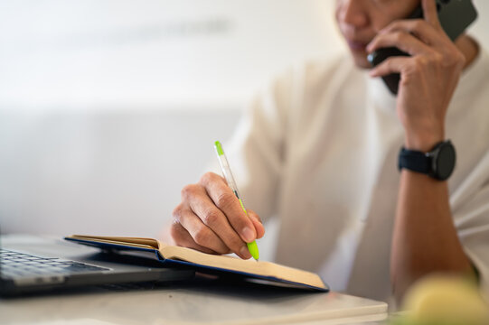 Asian man multitasking at a café table, talking on the phone while writing notes in a notebook beside a laptop, showing focus and productivity in a modern workspace environment.