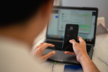 Asian man using a smartphone while sitting in front of a laptop with a messaging app on the screen, multitasking between phone and computer in a modern workspace with blurred background