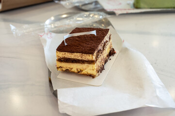 Close-up of a delicious slice of tiramisu cake with layers of cream and cocoa powder on top, served on white parchment paper over a marble table in a cozy café setting.