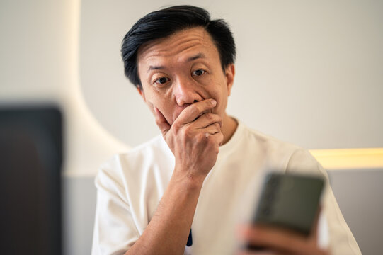 Asian man sitting at a café table, looking shocked while staring at his smartphone screen, reacting to surprising or unexpected news with an expressive face in a modern indoor workspace setting