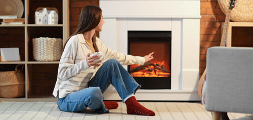 Young woman with cup of cocoa sitting near fireplace at home in evening