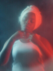 Conceptual portrait of young Black woman in white top and headwrap standing in the dark with closed...