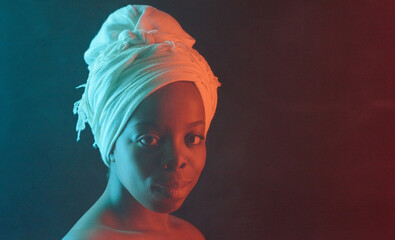Headshot portrait of young attractive African woman in white turban with naked shoulders smiling at camera on dark background, among fog