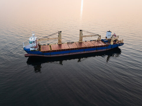 Aerial drone view of a geared bulk carrier, a dry cargo ship with cranes, sailing through the calm Baltic Sea at sunset.