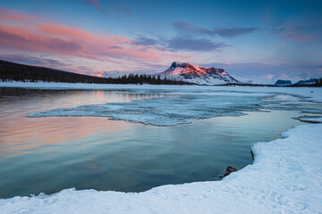 Winter sunrise reveals vibrant colors over frozen lake and snowy peaks at Sarek Nationalpark in...
