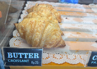 Golden butter croissants sit on a lace doily in a bakery display case, tempting customers with...