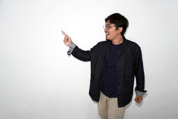 A cheerful asian man in glasses and a denim jacket points his finger to the side with a wide smile, enthusiastically presenting or directing attention. Copy space