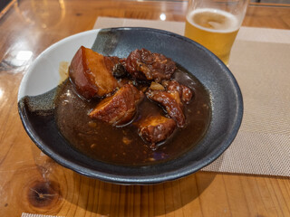 Tender, slow-cooked braised pork belly in a rich, dark savory sauce, presented in a unique bowl alongside a refreshing golden beer on a warm wooden dining table.