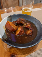 Delicious braised pork belly, glistening in a savory dark sauce, served in a rustic bowl on a wooden table. A refreshing glass of beer complements this hearty, home-cooked meal, perfect for a cozy