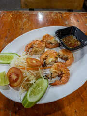 A mouth-watering plate of perfectly grilled jumbo prawns, served with a fresh side salad of shredded cabbage, cucumber, and tomato, alongside a zesty chili dipping sauce. A delicious and healthy