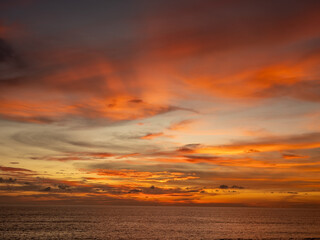 A breathtaking tropical sunset paints the expansive ocean horizon with vibrant streaks of fiery orange, golden yellow, and dramatic reddish-purple clouds. The tranquil water reflects the atmospheric
