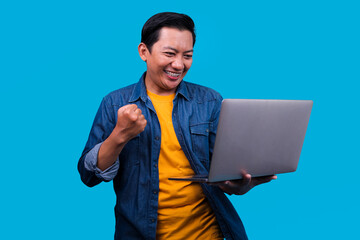 Asian Freelancer Man Holding Laptop While Raising Fist Celebrating Success Over Blue Studio Wall
