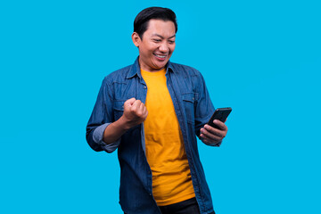 Asian Young Guy Raising Fist While Looking At Smartphone Over Blue Background