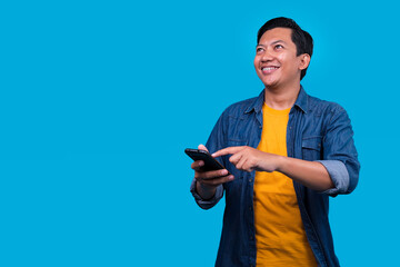 Smiling Asian Man Holding Mobile Phone While Looking Up to Empty Space Isolated on Blue Background