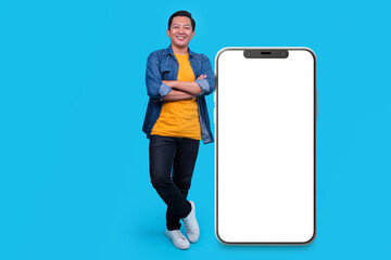 Full Length of Asian Man Standing Near Big Huge Blank Screen of Mobile Phone on Blue Studio