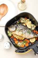 Cooked fish, lemon and vegetables in air fryer basket on white wooden table, flat lay