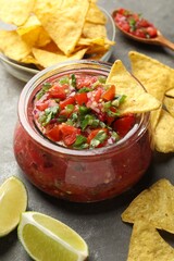 Tasty salsa in jar, lime and nachos on grey table, closeup
