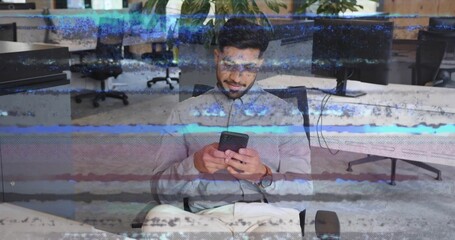 Indian office worker wearing glasses looking at smartphone sitting at desk in office, with monitors © vectorfusionart