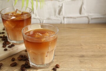 Tasty iced coffee with milk in glasses and beans on wooden table against light textured background, closeup. Space for text