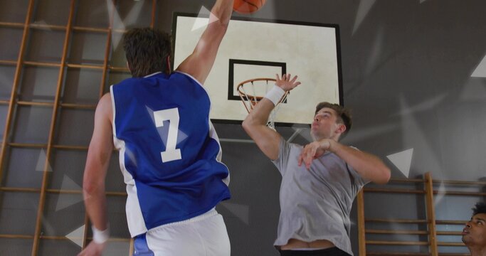 Leaping player wearing blue-white jersey shooting basketball toward hoop in gym, defender blocking - Powered by Adobe