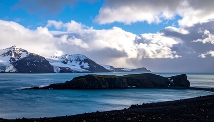Snowy mountains and a dark, rocky land mass meet the aqua sea beneath a cloudy sky with a sliver of sunshine