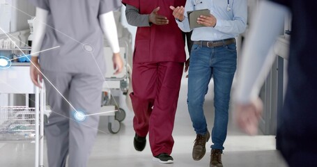 Walking nurse wearing burgundy scrubs, doctor with tablet sharing patient info in hospital corridor