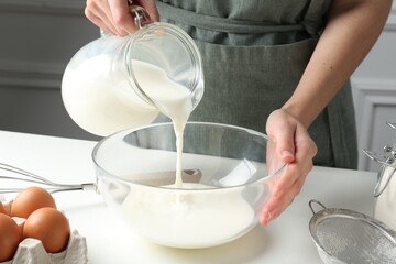 Making batter (liquid dough). Woman pouring milk into bowl at white table indoors, closeup