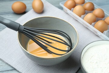 Beaten eggs in bowl, whisk and flour on table, closeup
