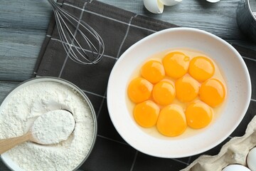 Raw eggs, yolks in bowl, flour and whisk on wooden table, flat lay