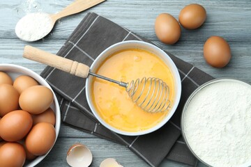 Beaten eggs in bowl, whisk and flour on wooden table, flat lay