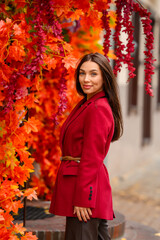 Portrait of a young woman with leaves in an autumn park. A stylish girl in a burgundy jacket strolls outdoors. Autumn fashion.