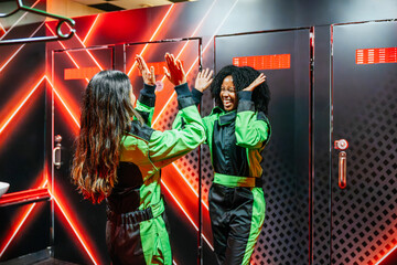 Two women high fiving in racing suits, confidently preparing for a fun go karting experience inside a changing room © Koldo_Studio