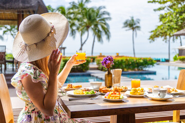 Woman in sun hat enjoys tropical breakfast by resort pool. Fresh juice, fruit plate, palm trees...