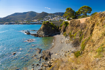 Fototapeta premium Secluded cove at l’Enbarril beach along the coastal path from Llançà to Port de la Selva, with cliffs, turquoise water, and a hillside village in the background.