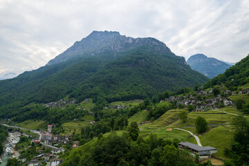 Aerial view of the quaint village nestled amid lush green slopes, shadowed by the imposing, rocky peak of a mountain, evoking a sense of serene isolation, Lavertezzo, Ticino, Switzerland.